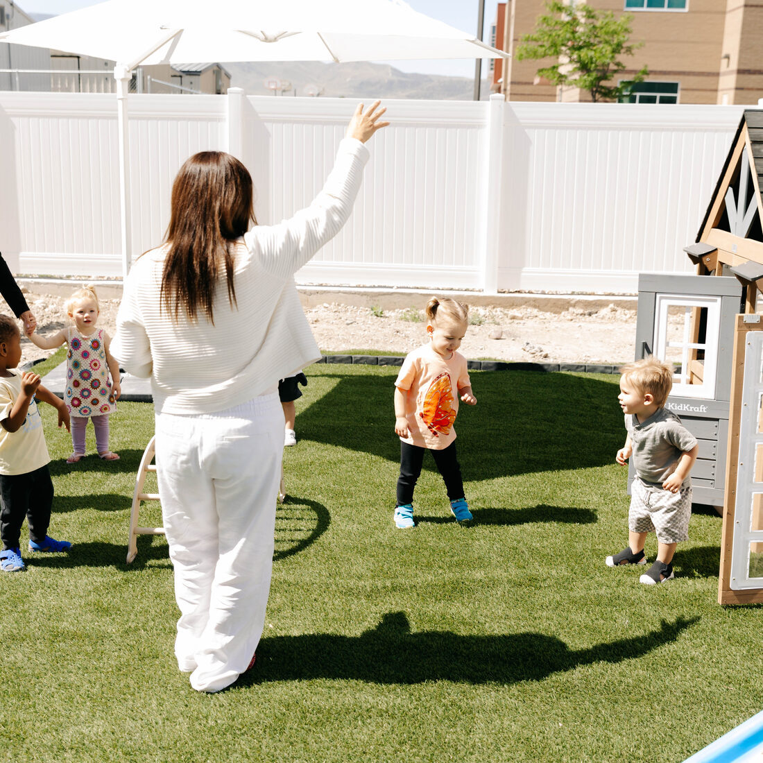 A teacher outside on the turf with a group of children, one arm raised mid-game.